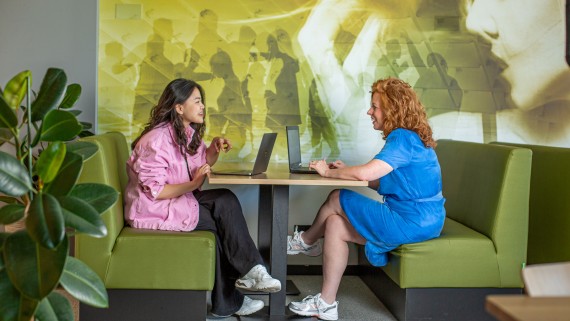 Docent en student zitten met laptops aan een tafel 