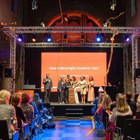 Een publiek luistert naar een presentatie in een grote zaal