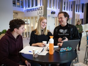 Een docent praat met twee studenten aan een statafel