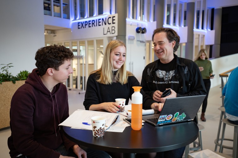 Een docent praat met twee studenten aan een statafel