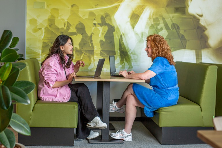 Docent en student zitten met laptops aan een tafel 