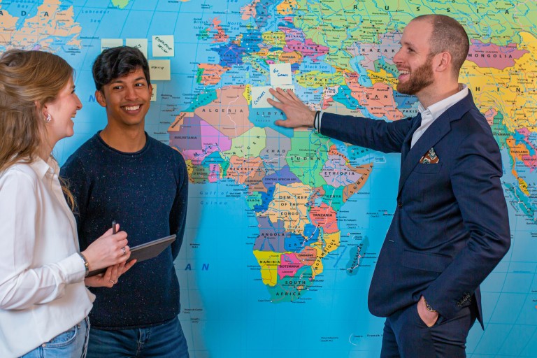 Een docent en twee studenten staan voor een wereldkaart
