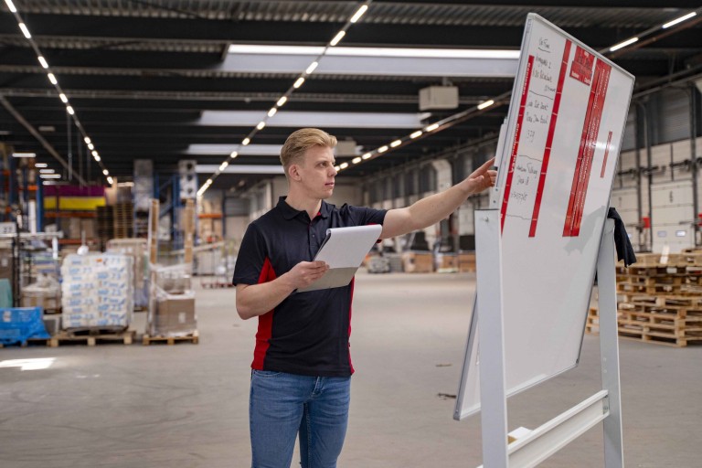 Een man in een magazijn wijst iets aan op een whitebord