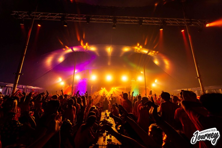 Feestende menigte met armen omhoog onder felle podiumverlichting in een festivaltent.