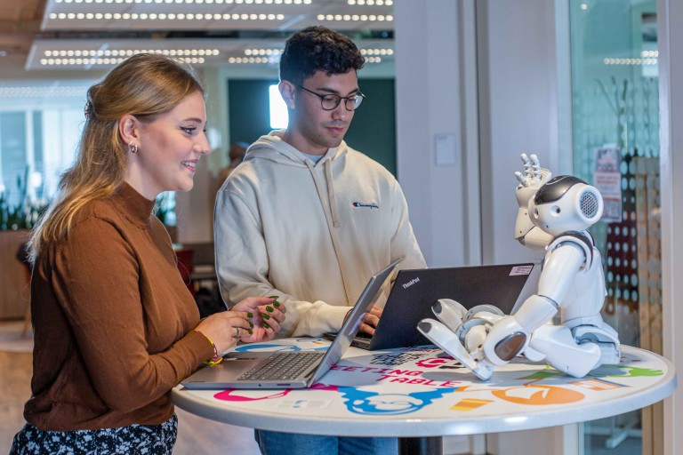 Twee studenten werken met laptops aan een sta-tafel naast een mensachtige robot