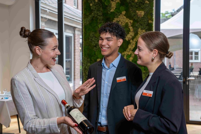 Studenten in pak presenteren een wijnfles aan een lachende gastvrouw in een restaurant.