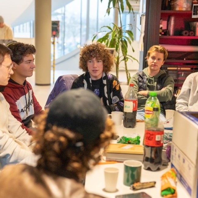 Zeven studenten praten aan een rommelige tafel